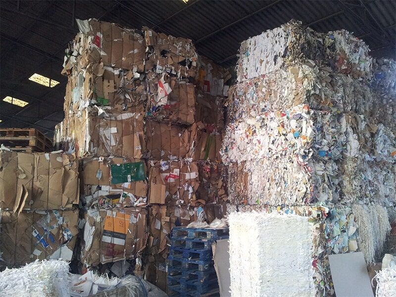 Pilhas de papelão prensado e fardos de papel em um armazém, alguns sobre paletes de madeira.