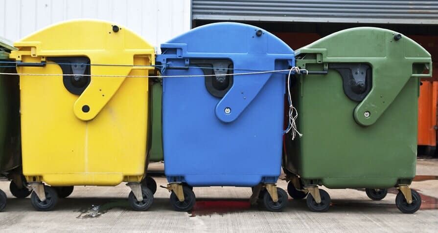 Lixeiras de reciclagem amarelas, azuis e verdes sobre rodas, enfileiradas ao ar livre.