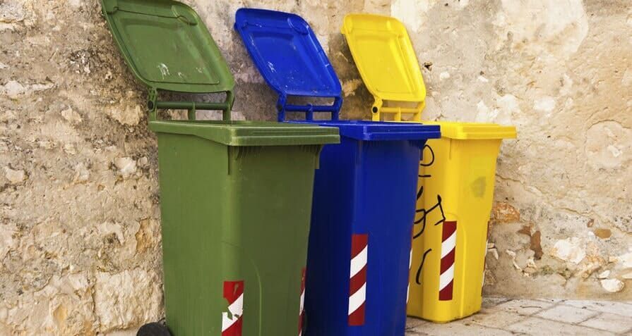 Lixeiras de reciclagem verdes, azuis e amarelas com as tampas abertas estão encostadas em uma parede de pedra.