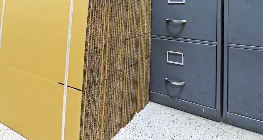 Pilha de folhas de papelão presas com fita adesiva branca ao lado de arquivos cinzentos em um piso salpicado.