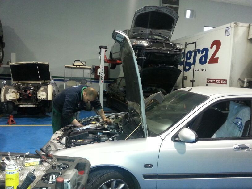 Un mecánico trabaja en un coche plateado en un taller. Hay varios vehículos con el capó abierto.