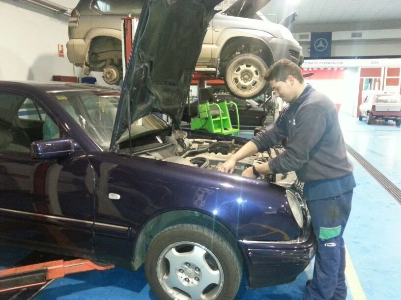 Un mecánico con overol azul trabaja en un coche azul en un taller. Al fondo, otro vehículo es elevado con una grúa.