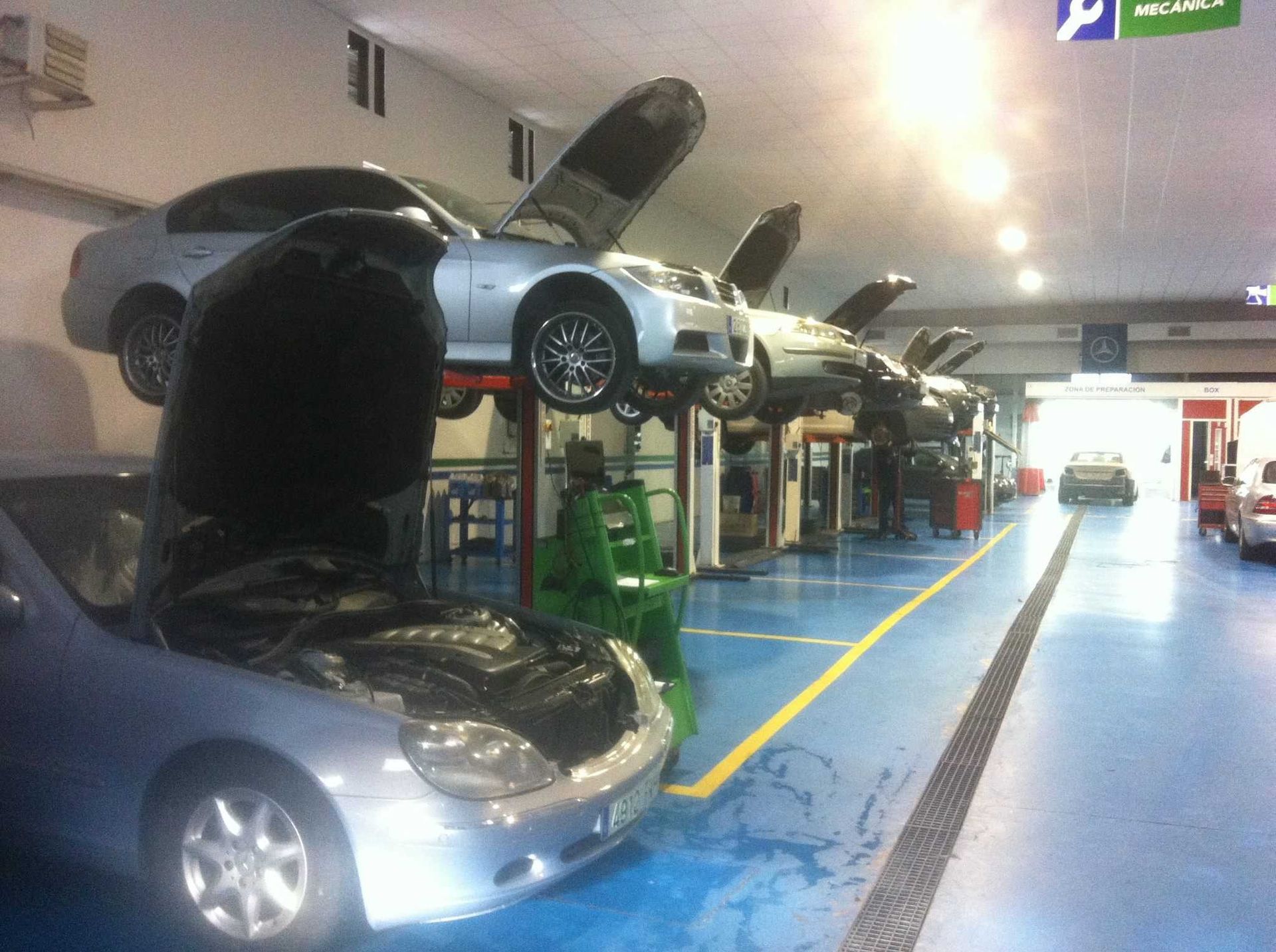 Autos subidos en elevadores en un taller mecánico, con el capó abierto. Se ven el suelo azul, una línea amarilla y una caja de herramientas verde.