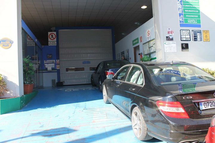 Coches alineados en una inspección técnica de vehículos. Un coche negro está en primer plano, esperando entrar en un taller.