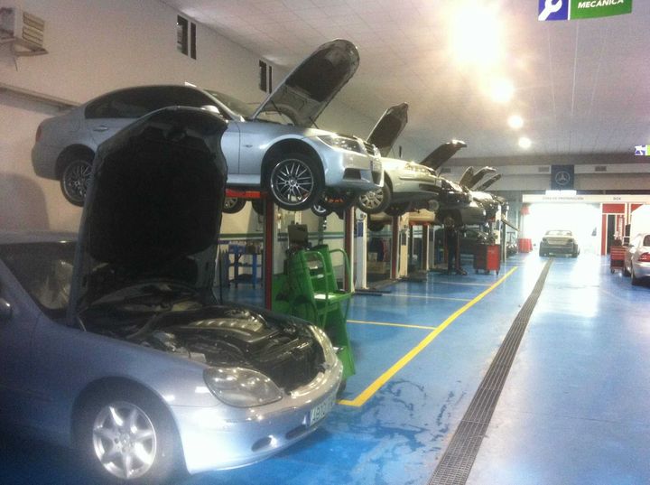 Coches en elevadores en un concurrido taller mecánico, con el capó abierto. Tonos azules y grises dominan el interior.