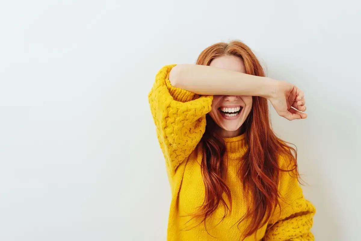 Una mujer pelirroja se ríe, tapándose los ojos con el brazo, vistiendo un suéter amarillo, contra una pared blanca.