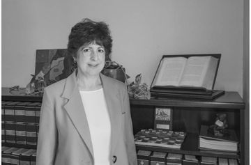 Una mujer con traje está parada frente a una estantería con una Biblia encima.