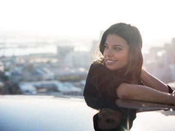 Mujer sonriendo junto a un coche al atardecer, con el horizonte de la ciudad borroso al fondo.