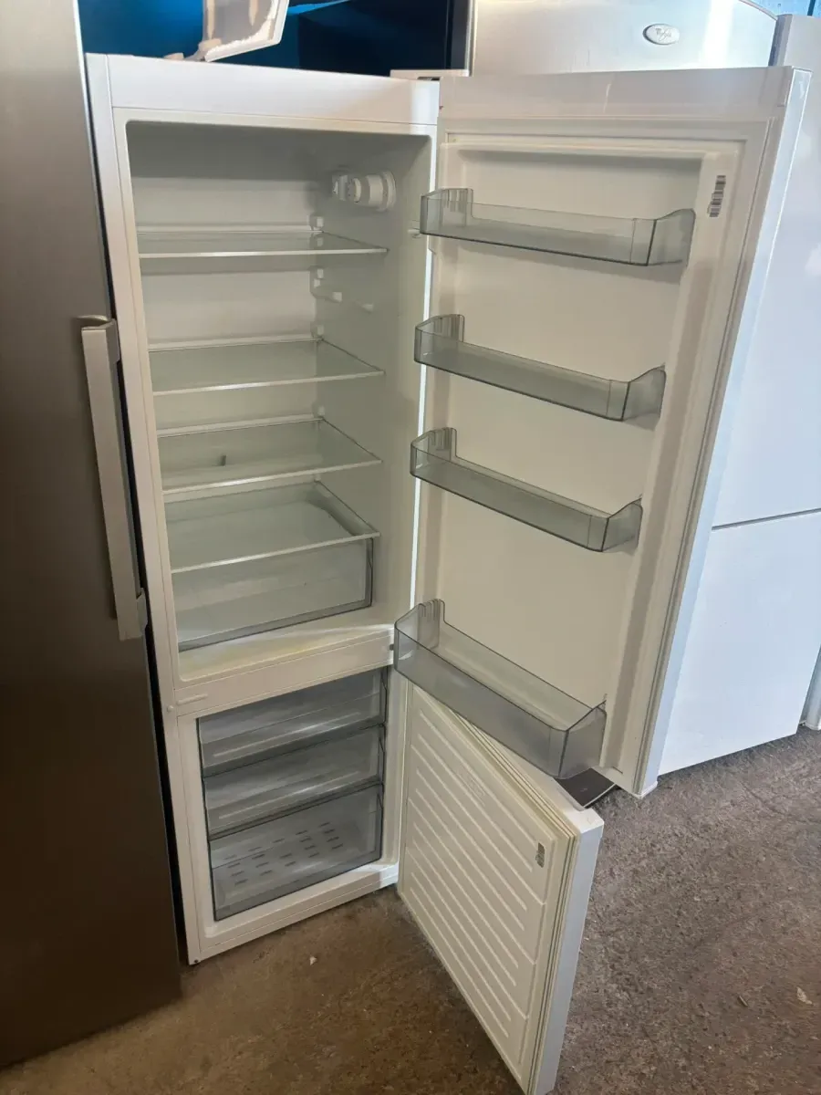 Congelador vertical abierto con estantes y compartimentos de la puerta vacíos, panel frontal abierto en una cocina