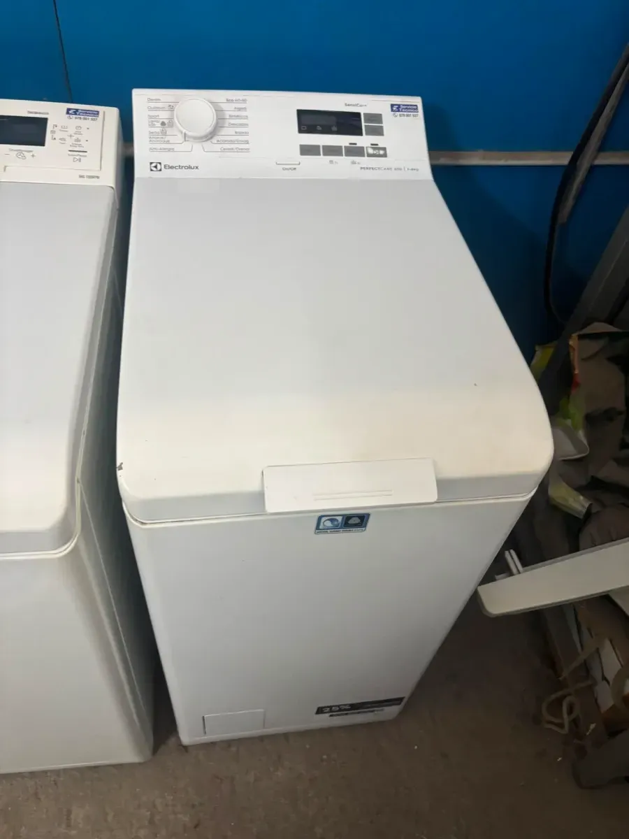 Lavadora de carga superior blanca en un área de servicio, con un panel de control y una pared azul detrás.
