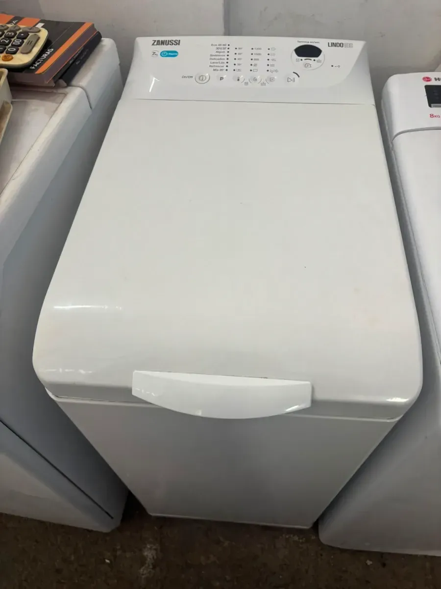 Lavadora blanca de carga superior situada entre dos electrodomésticos, con panel de control en la parte superior.