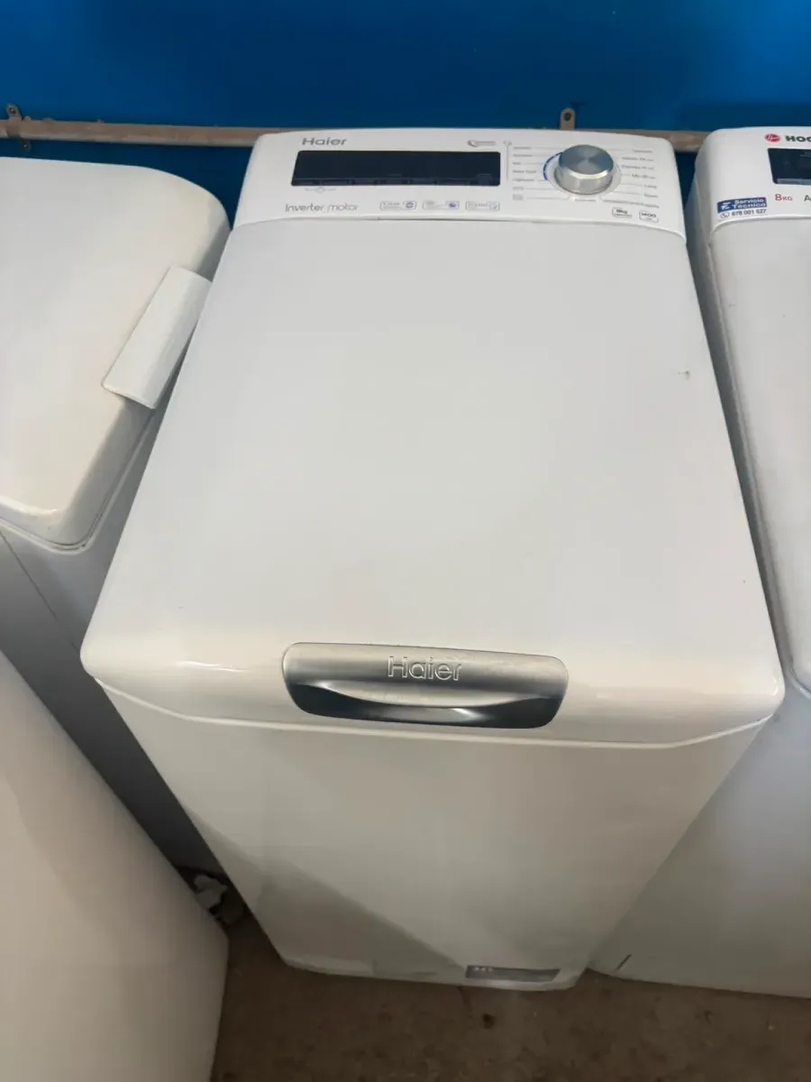 Lavadora blanca de carga superior en una zona de lavandería, con panel de control y tirador plateado.
