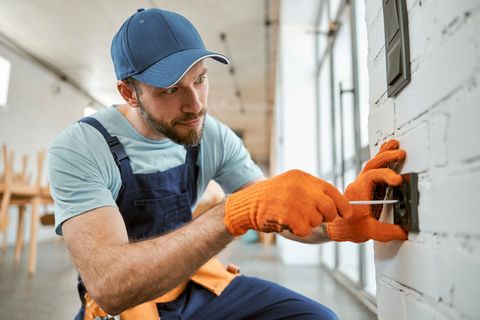 Un électricien en combinaison avec des gants de travail utilise un tournevis sur une prise murale.