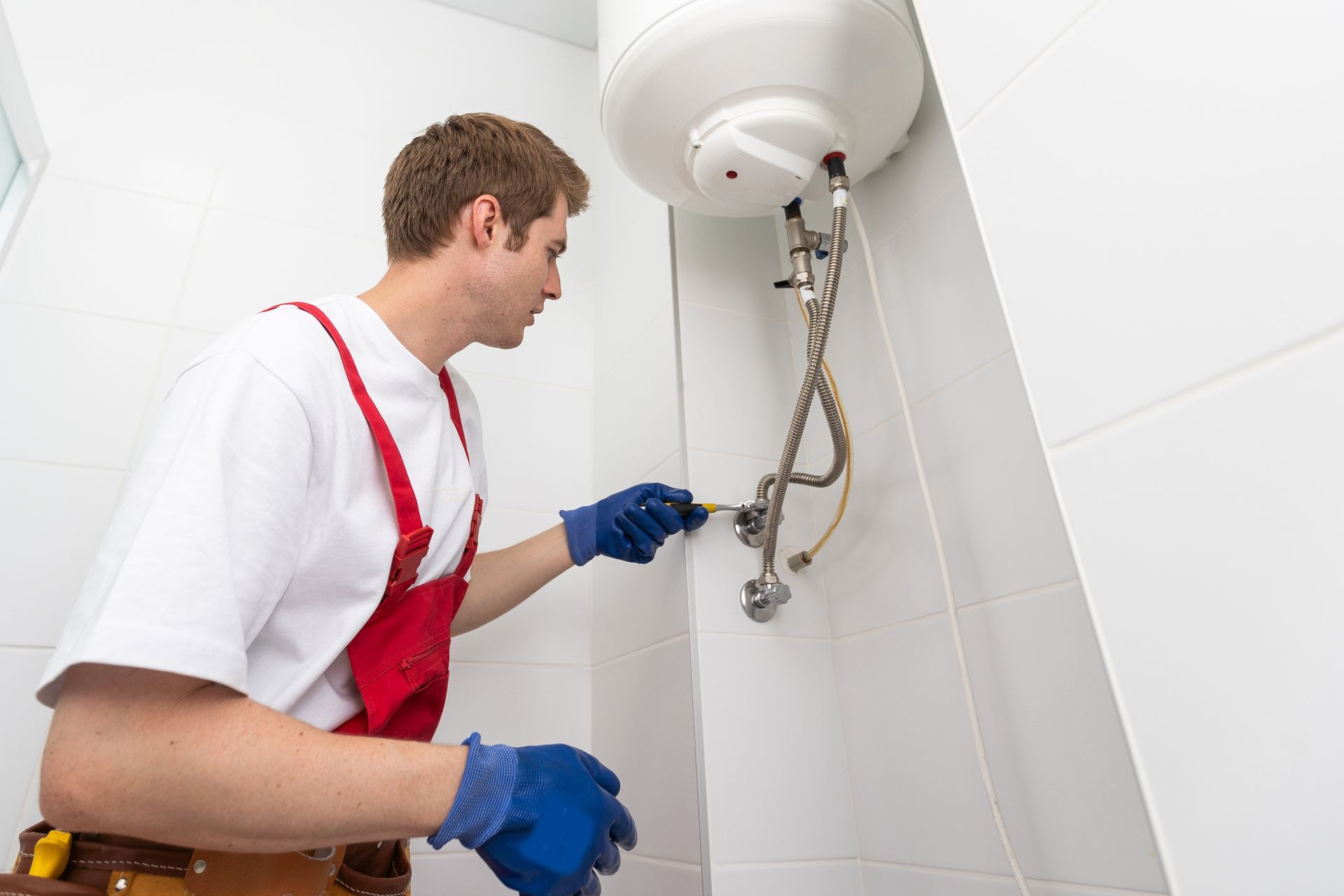 Un plombier en salopette rouge et gants bleus travaille sur un chauffe-eau dans une pièce carrelée de blanc.