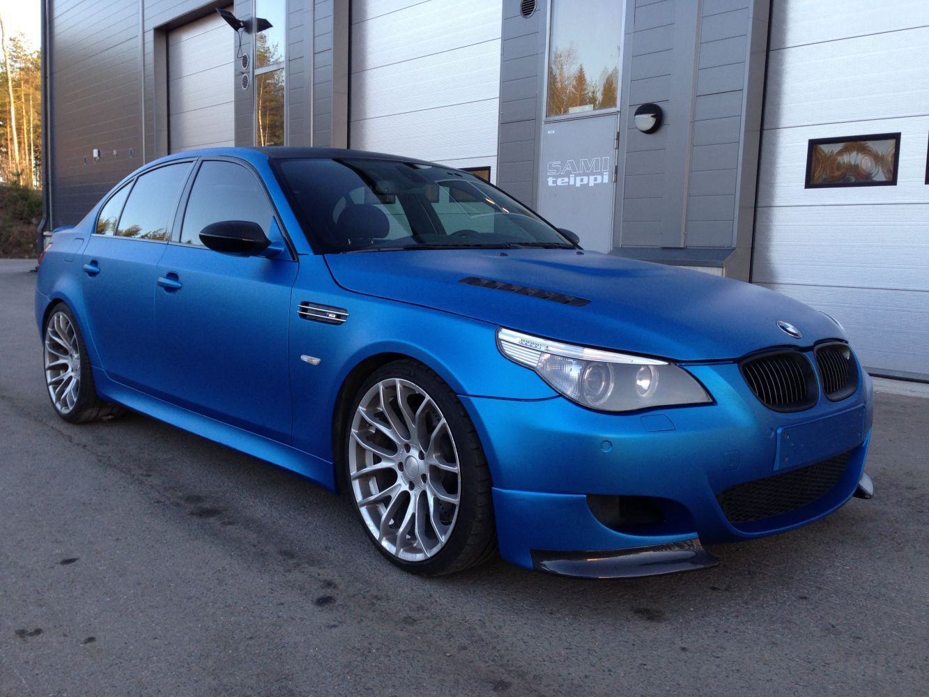 Sininen BMW M5 -sedan pysäköitynä rakennuksen eteen, jossa on autotallin ovi.