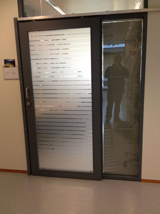 Ovi, jossa on himmeä lasi ja vaakasuoria raitoja, kaihtimilla varustetun ikkunan vieressä rakennuksessa. Ihminen heijastuu lasista.