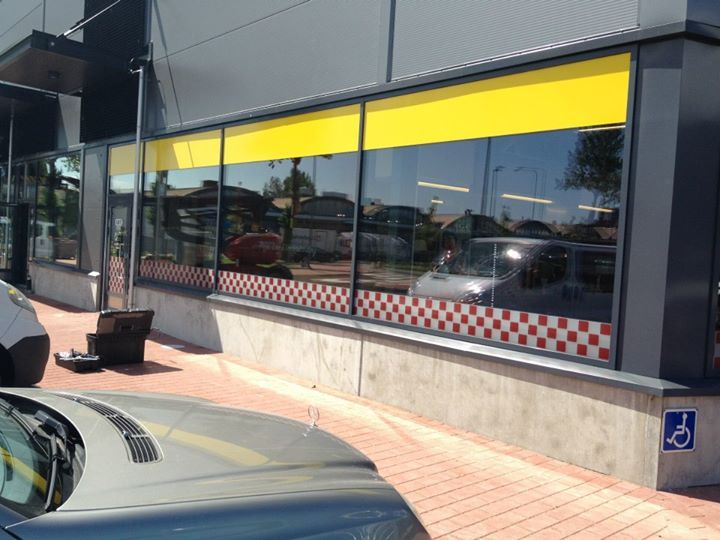 Rakennuksen ulkokuva, jossa on suuret ikkunat, punainen ja valkoinen ruutunauha ja keltainen markiisi. Mukana on myös invapysäköintisymboli.