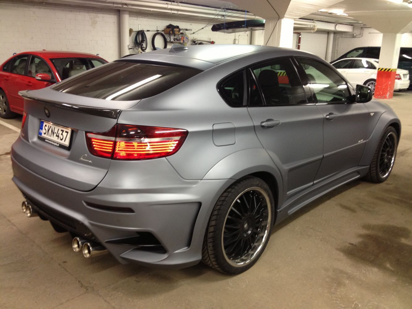 Harmaa muunneltu BMW X6 -auto maanalaisessa parkkihallissa, jossa on mustat vanteet ja rekisterikilpi.