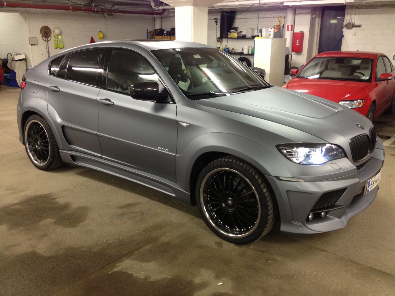 Harmaa BMW X6 mustilla vanteilla pysäköitynä autotalliin, punainen auto taustalla.