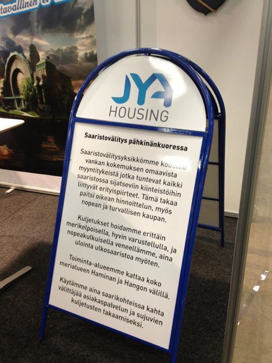 JYA Housingin A-mallinen kyltti, sinivalkoinen, suomenkielisellä tekstillä, sisätiloissa tapahtumassa.