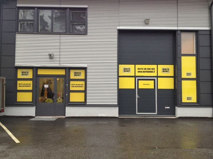 Liikerakennuksen ulkopinta harmaalla ja keltaisella julkisivulla, ovella, autotallilla ja ikkunoilla.