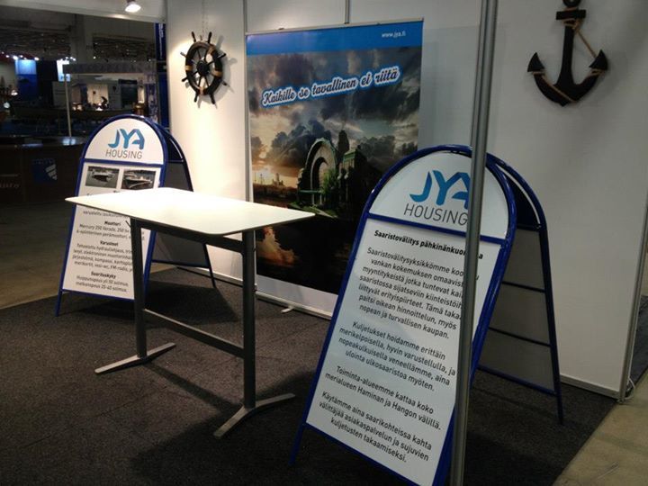 Koju, jossa on kylttejä, pöytä ja ankkurikoristeita. Sinivalkoinen kyltti tekstillä, mahdollisesti venenäyttelyssä.