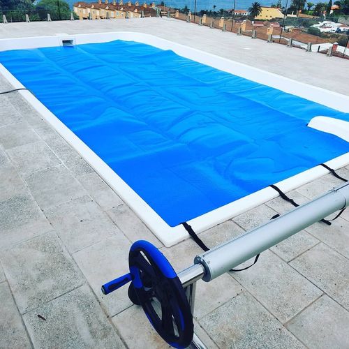 Una lona azul para piscina, extendida sobre una piscina en la azotea con un rodillo metálico sobre una terraza embaldosada