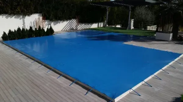 Piscina exterior cubierta con una lona azul, rodeada por una terraza de madera y árboles.