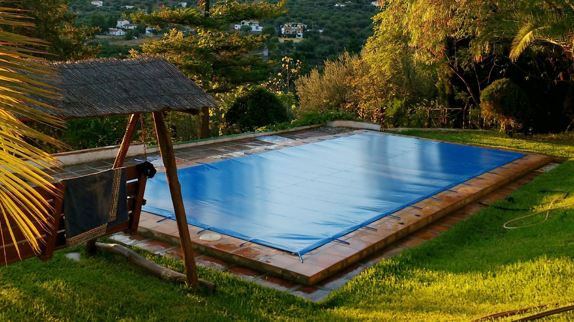 Una cubierta azul para la piscina junto a un columpio de madera en el patio trasero de un frondoso jardín.