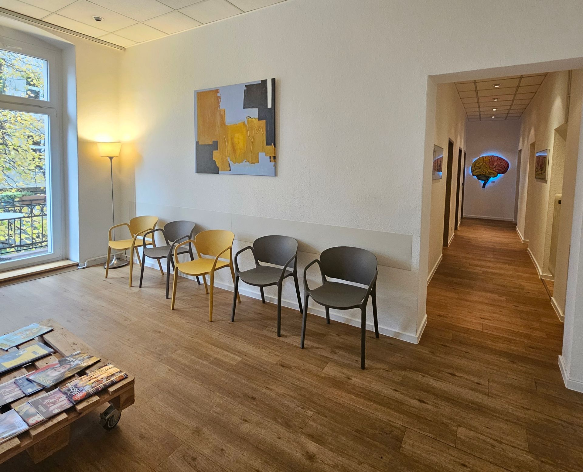 Wartezimmer mit Stühlen, Kunstwerken und einem Flur, der zu einem Bild mit Gehirnmotiv führt.