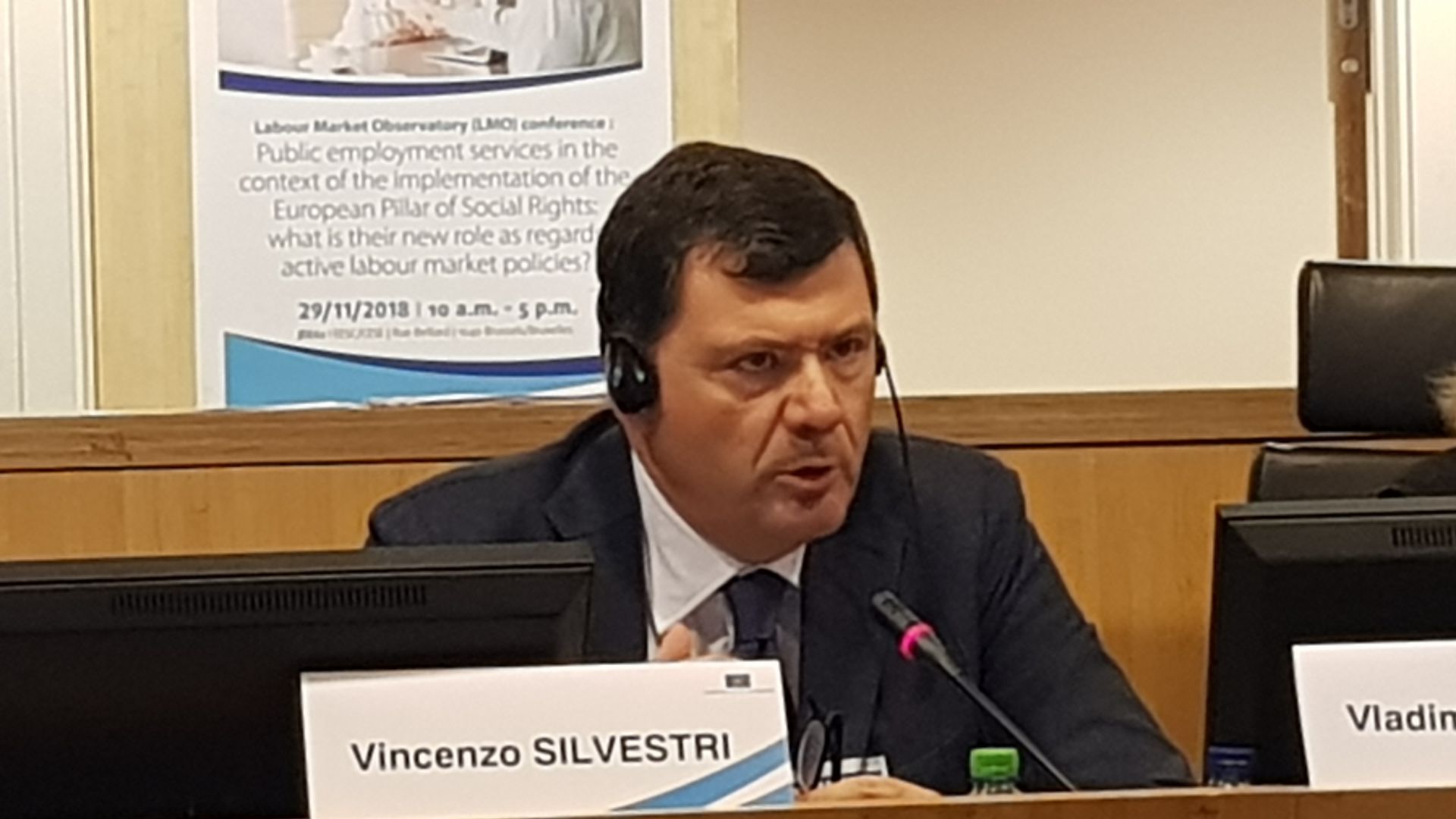 Un uomo in giacca e cravatta è seduto a un tavolo davanti a un microfono.