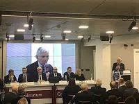 Un gruppo di persone è seduto davanti a un grande schermo mentre un uomo tiene un discorso.