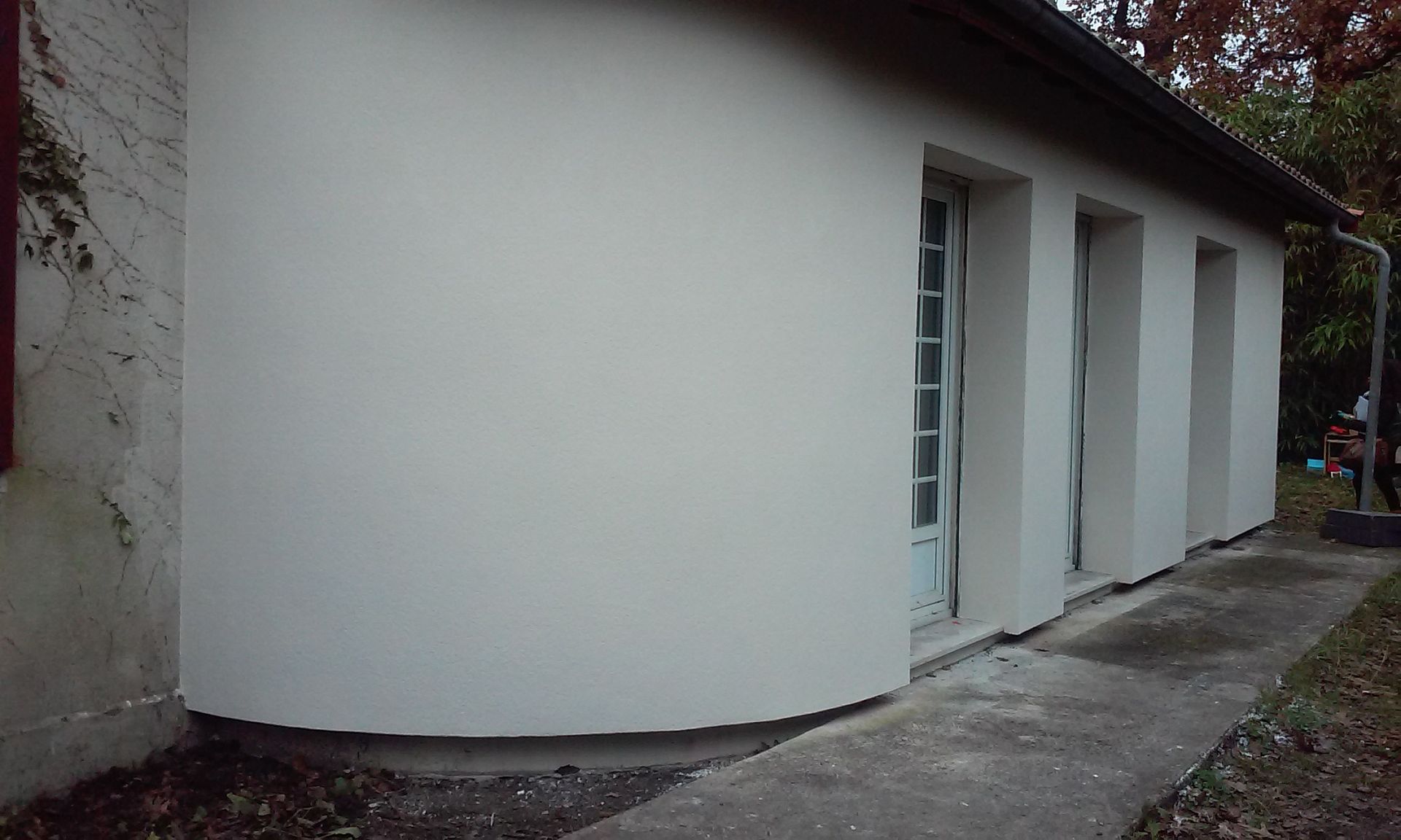 Un mur extérieur blanc incurvé, percé de trois portes vitrées, se détache sur une allée en béton.
