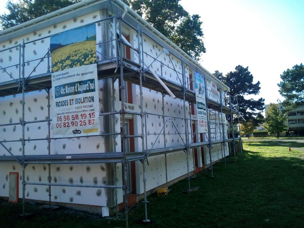 Un bâtiment en rénovation, avec des panneaux d'isolation blancs sur sa façade, entouré d'échafaudages sur une pelouse.