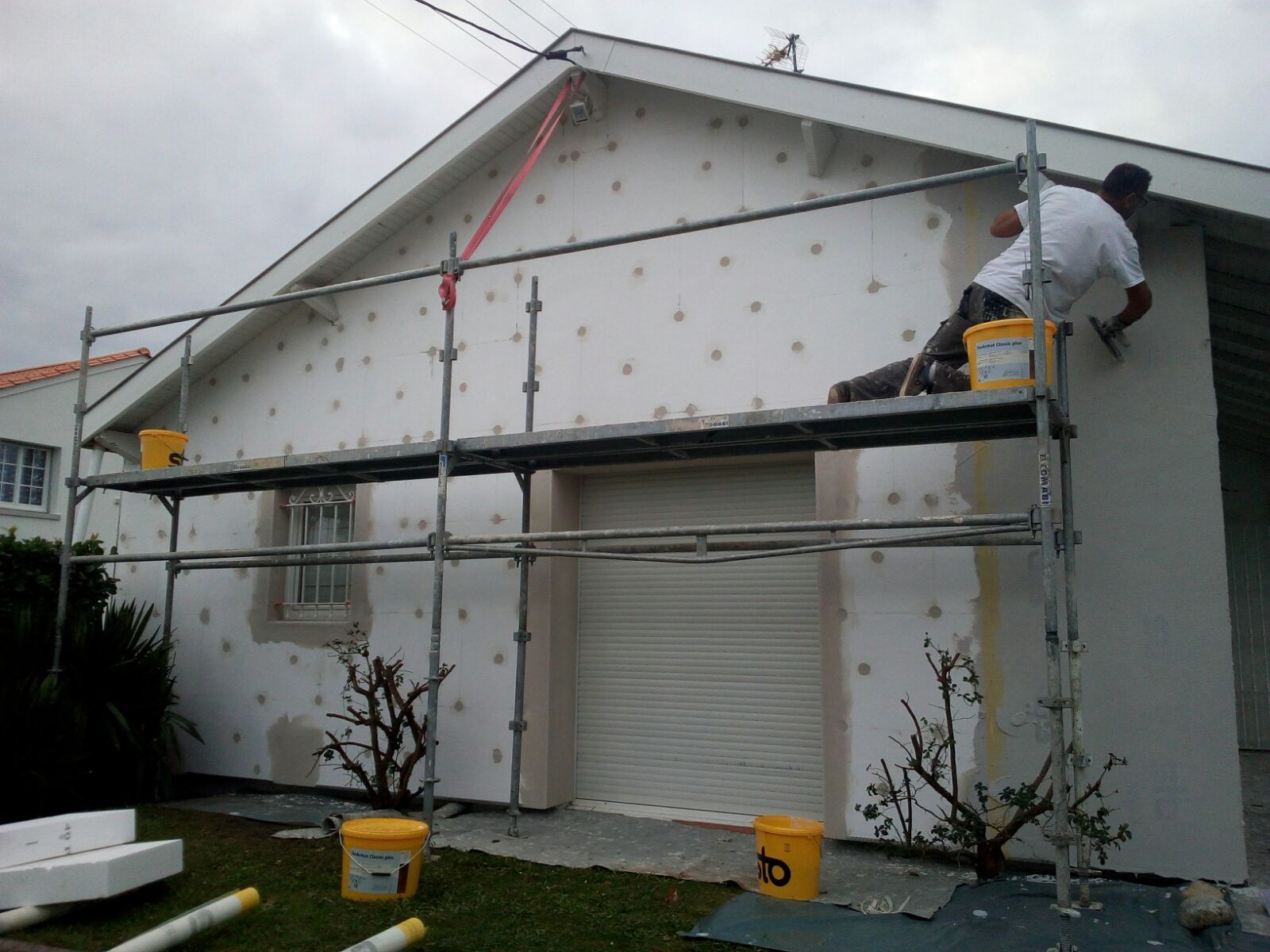 Un ouvrier, juché sur un échafaudage, applique un enduit sur le mur extérieur d'une maison recouverte de panneaux isolants.