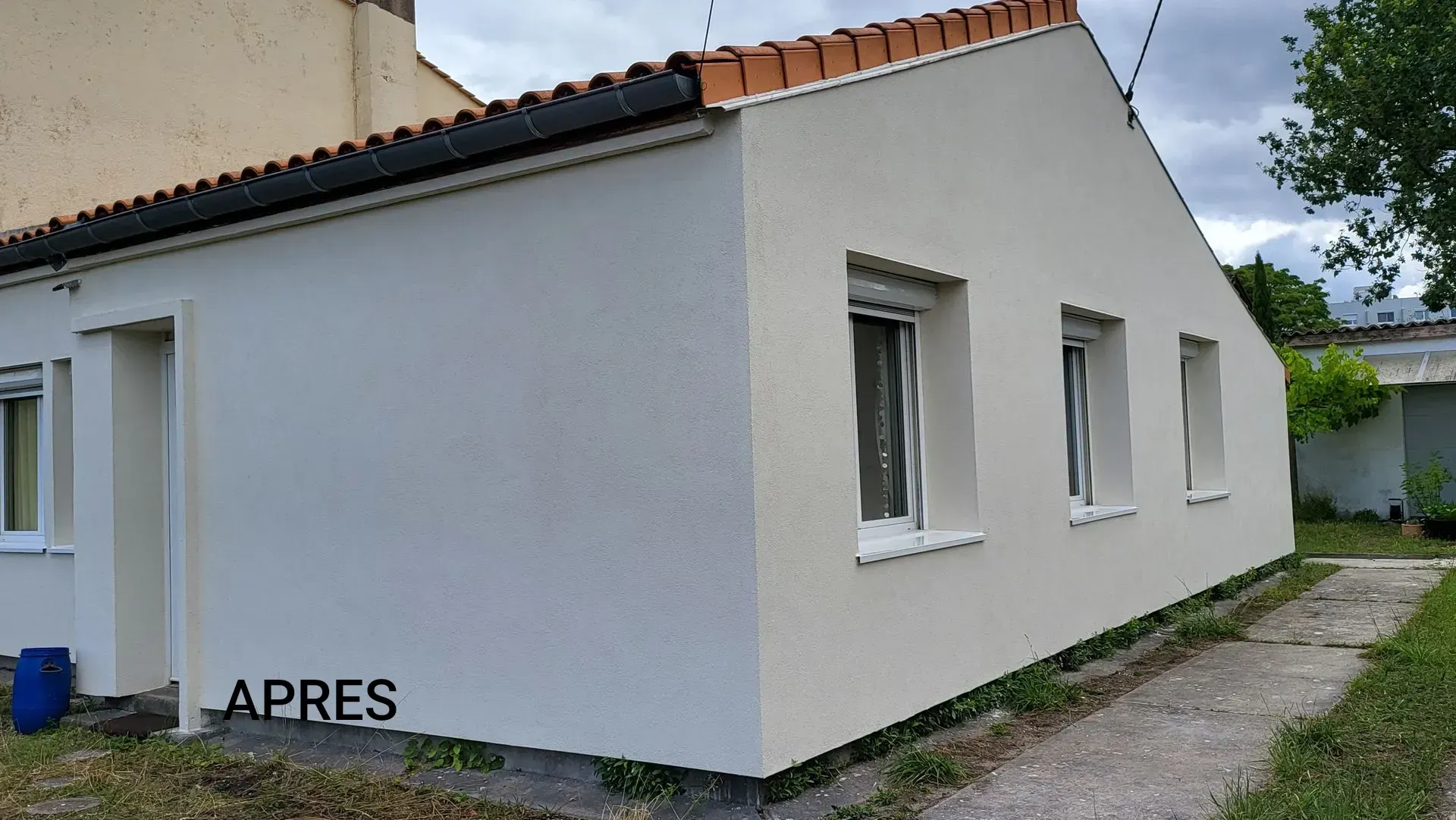 Façade d'une maison de plain-pied blanche, fraîchement enduite, avec un toit de tuiles, vue de côté par temps couvert.