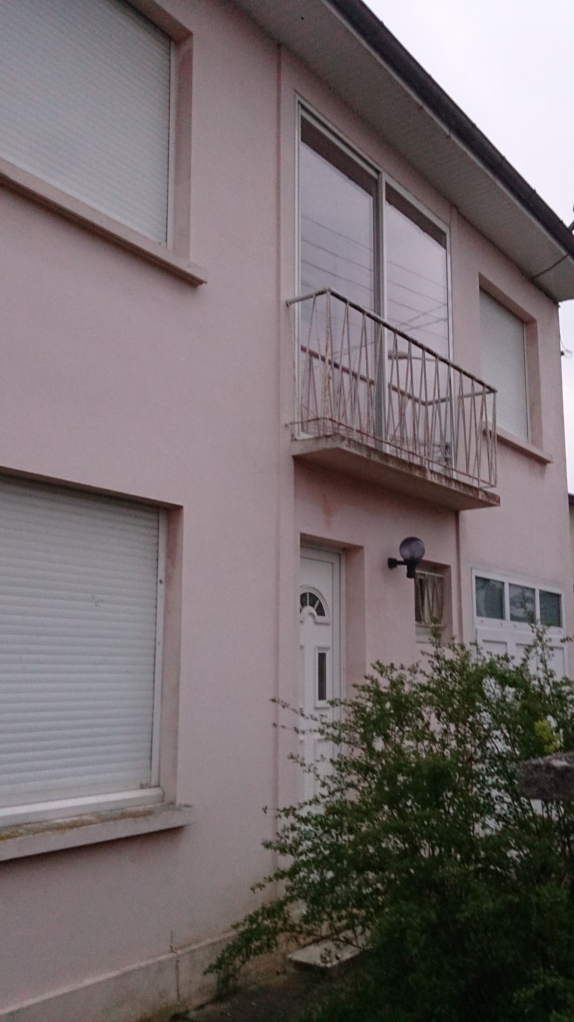 Une maison rose pâle de deux étages, avec un petit balcon en métal, des volets fermés et une entrée principale.