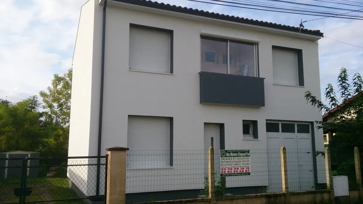 Une maison d'habitation blanche de deux étages avec un balcon gris foncé et un garage, derrière une clôture en fil de fer.