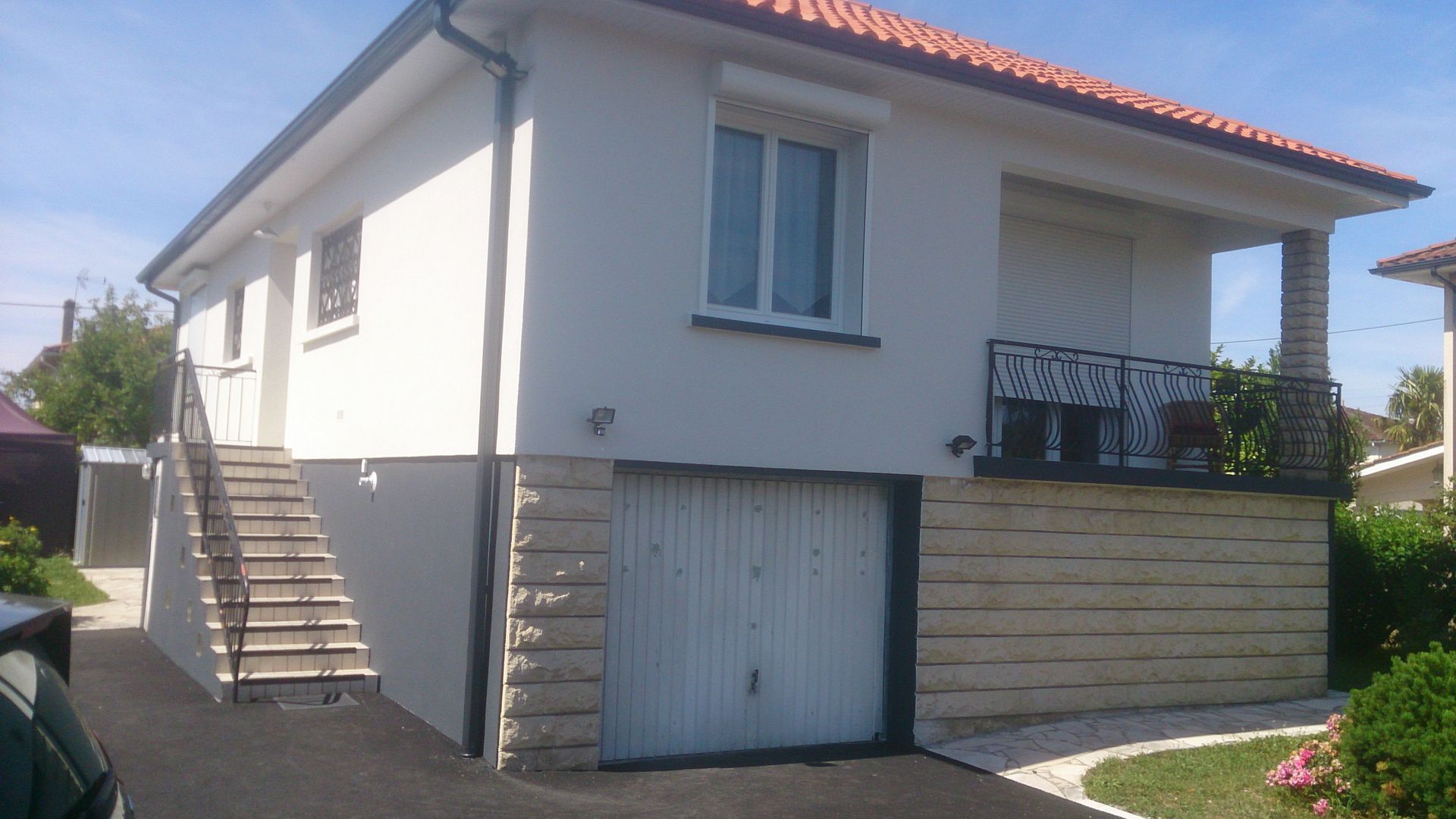 Maison à deux étages avec une façade blanche, un garage, un toit de tuiles, un escalier latéral et un balcon avec rambarde.