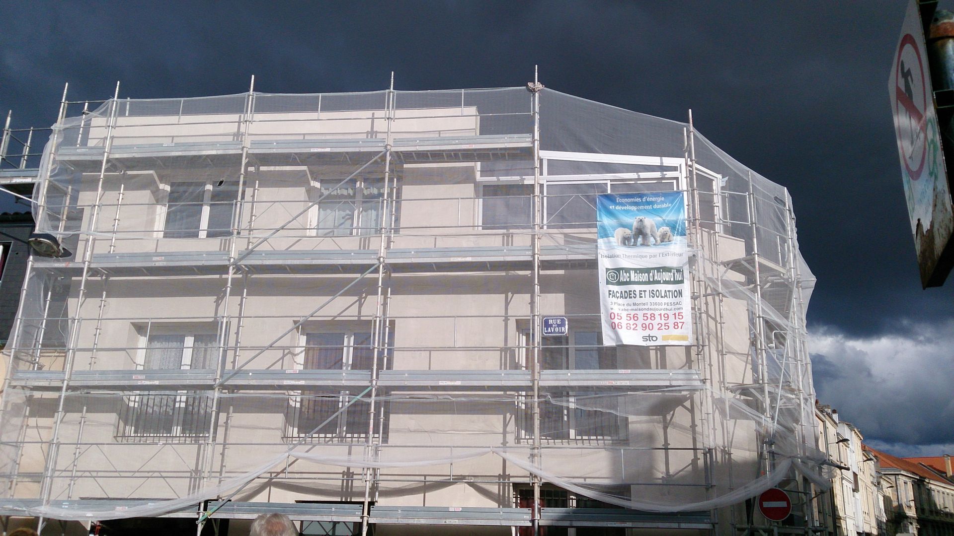 Un bâtiment clair sous un ciel sombre et orageux, entièrement entouré d'échafaudages de chantier et de filets de protection.