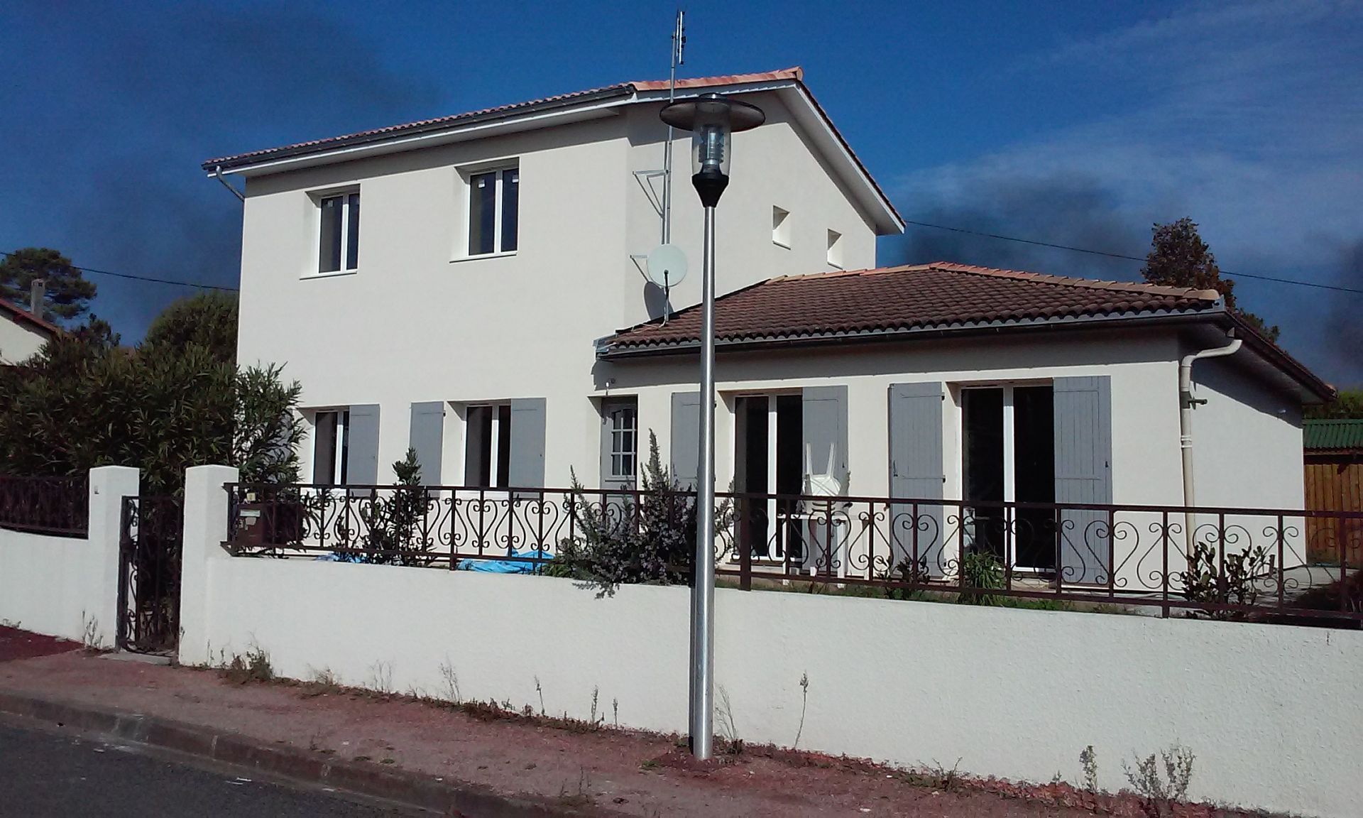 Une maison blanche à deux étages, avec des volets gris et un toit de tuiles, entourée d'un muret et d'une clôture métallique.