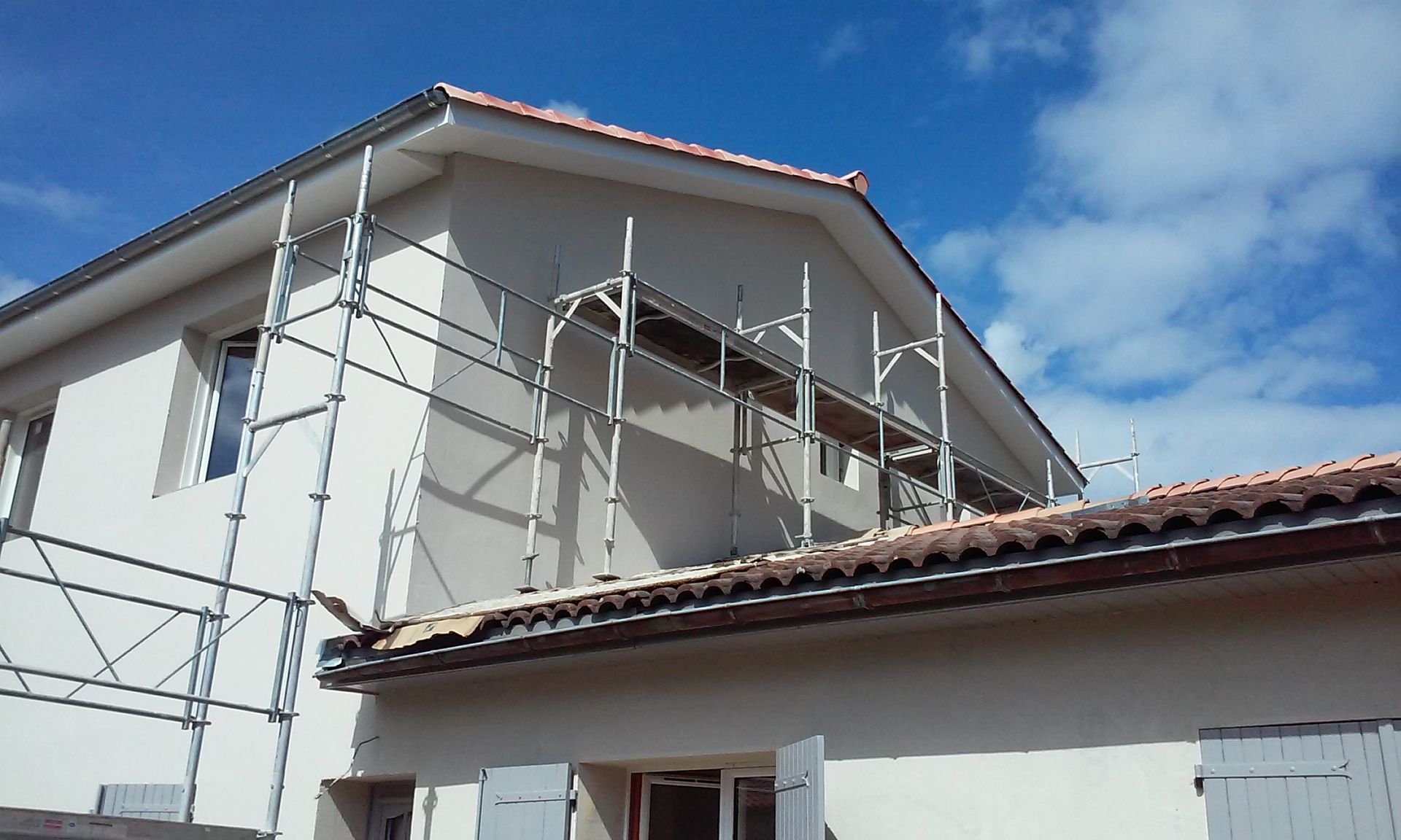 Échafaudages dressés contre le côté d'une maison beige à deux étages, sous un ciel bleu limpide.