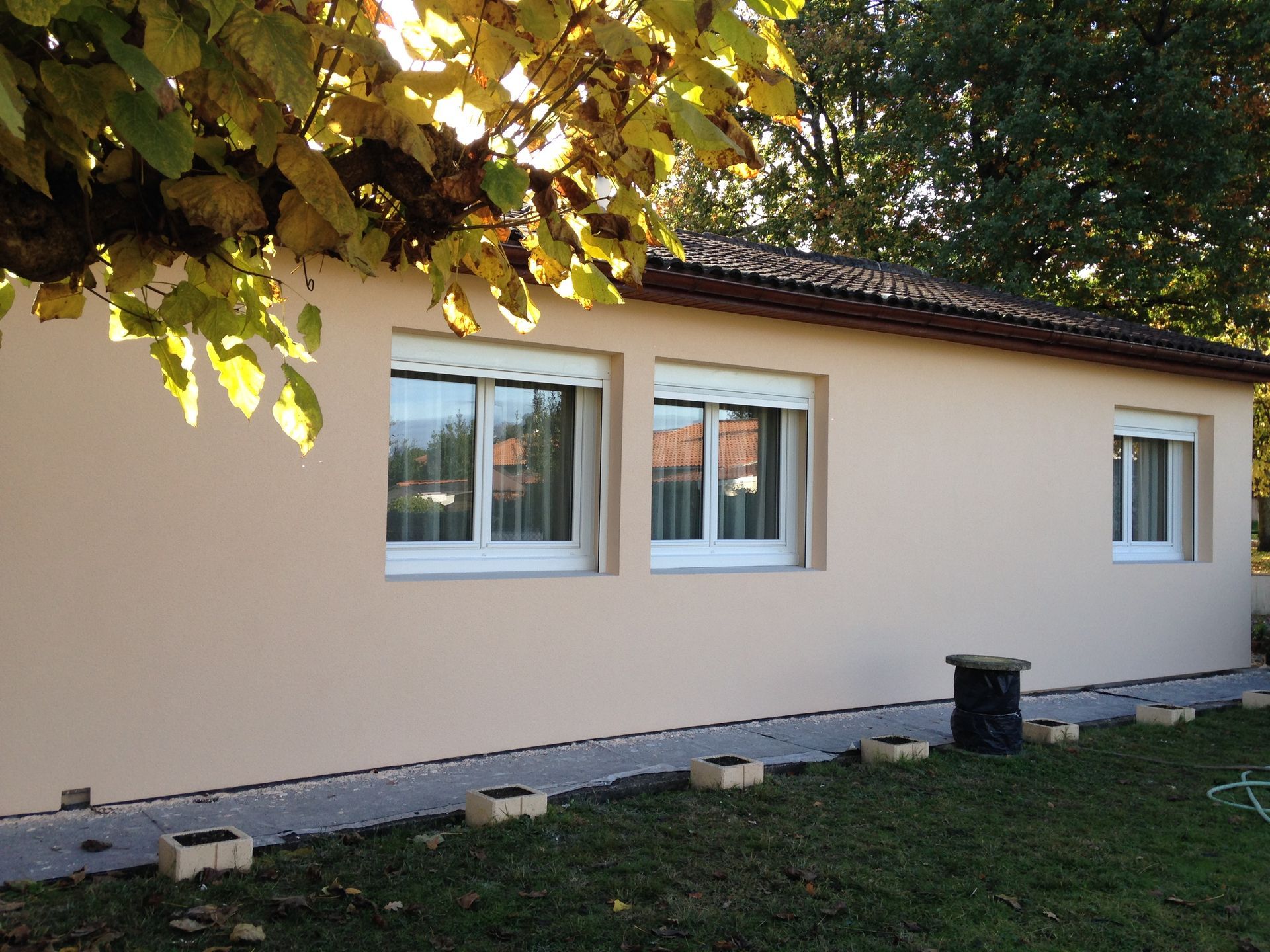 Maison de plain-pied, de couleur beige clair, avec trois fenêtres, un toit de tuiles et une allée en pierre dans un jardin arboré.