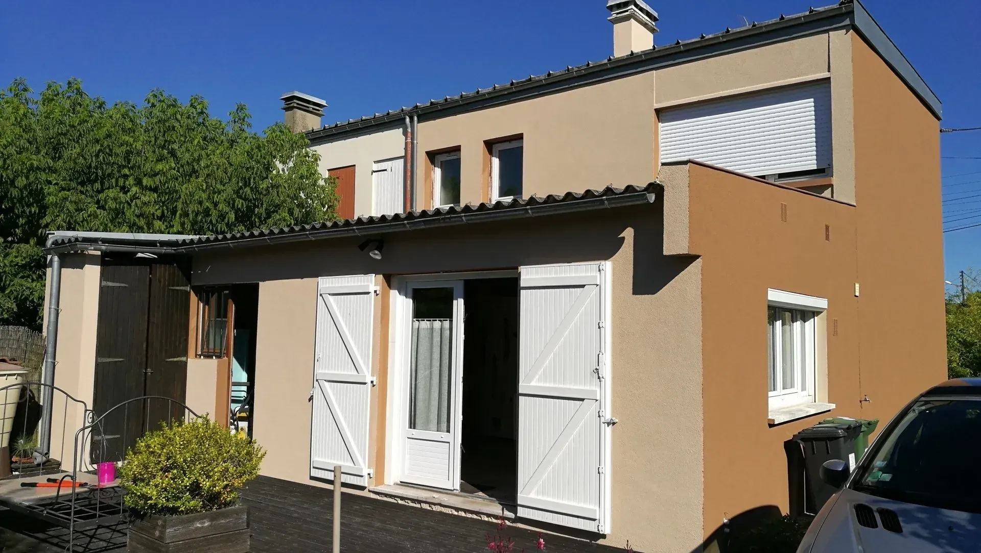 Une maison beige de deux étages avec des volets blancs, un toit sombre et une cour pavée devant, sous un ciel bleu clair.