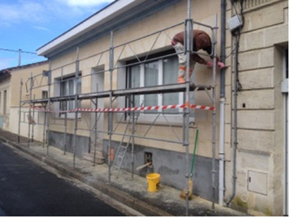 Une personne sur un échafaudage travaille sur le mur extérieur d'une maison en pierre beige, en bordure de rue.