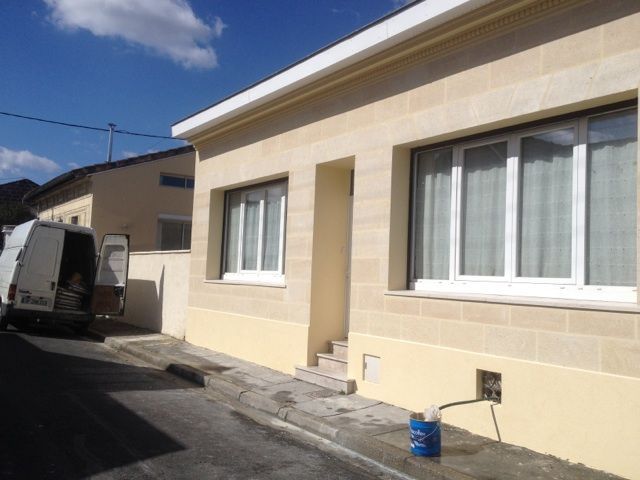 Maison de plain-pied en pierre claire, avec deux fenêtres, une entrée en retrait et une camionnette garée dans l'allée.