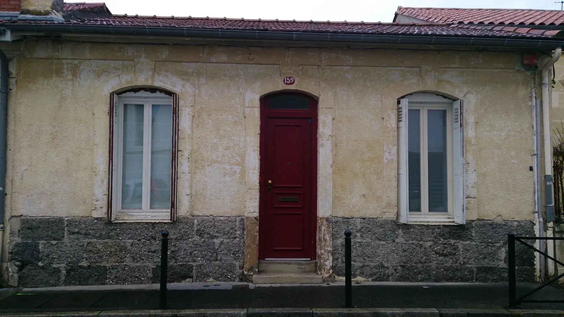 Une façade de maison avec une porte rouge flanquée de deux fenêtres, avant ravalement.