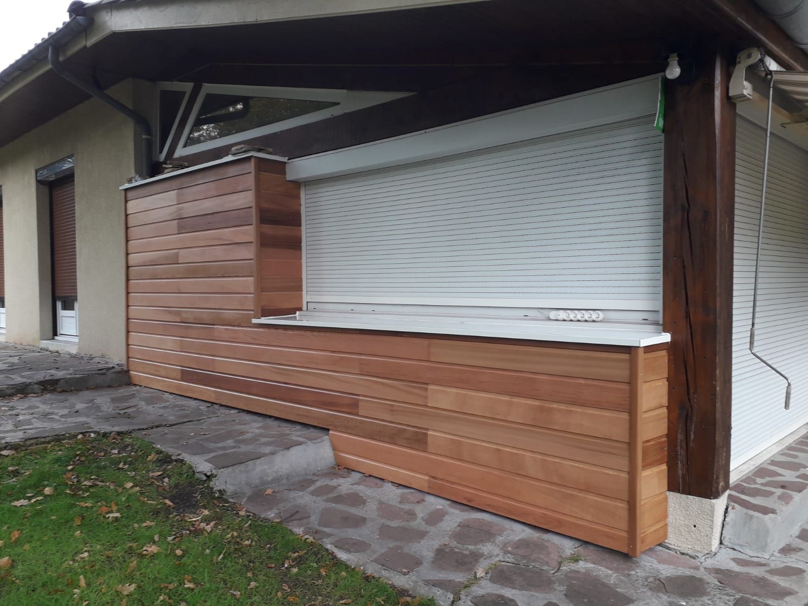 Un mur recouvert de bardage horizontal en bois et de volets roulants blancs, donnant sur une terrasse en pierre.