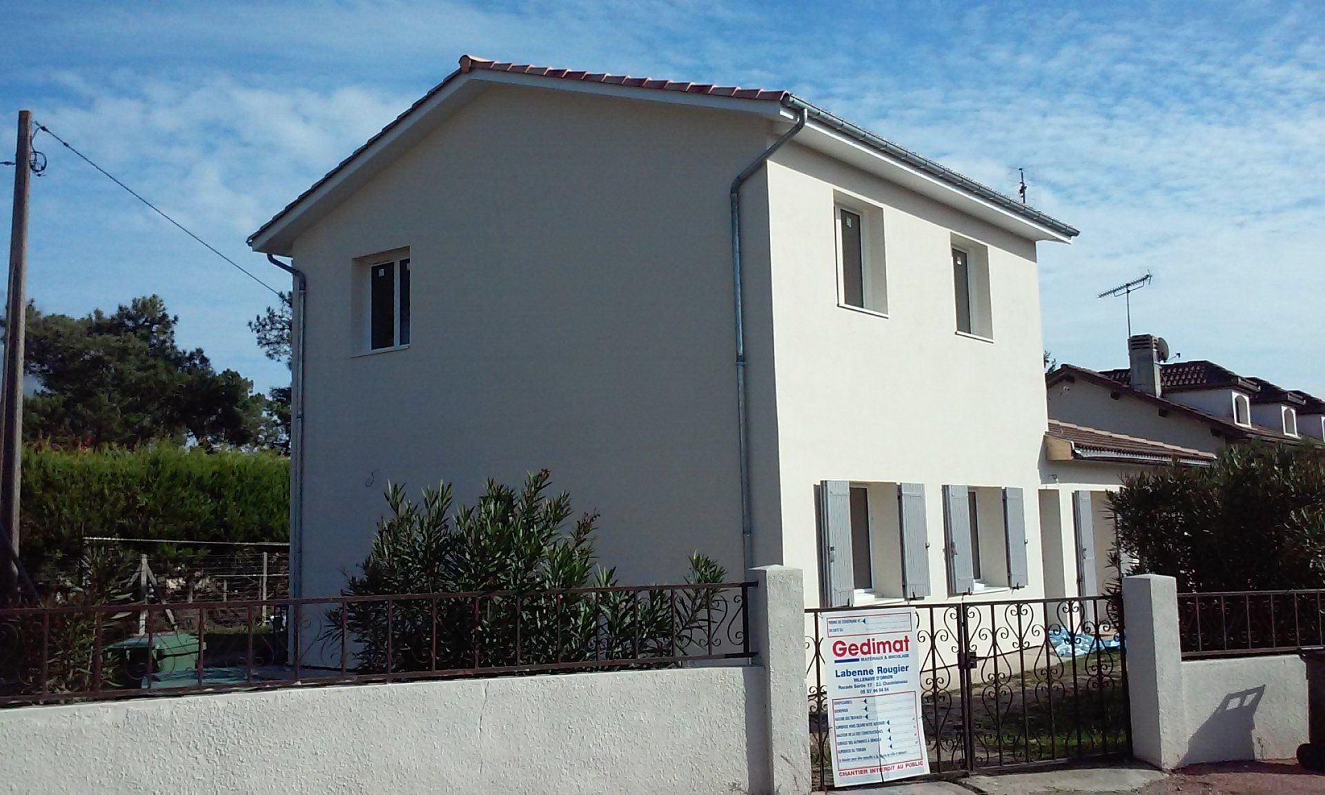 Une maison blanche à deux étages, aux volets clairs, se dresse derrière un muret de pierre, sous un ciel bleu.