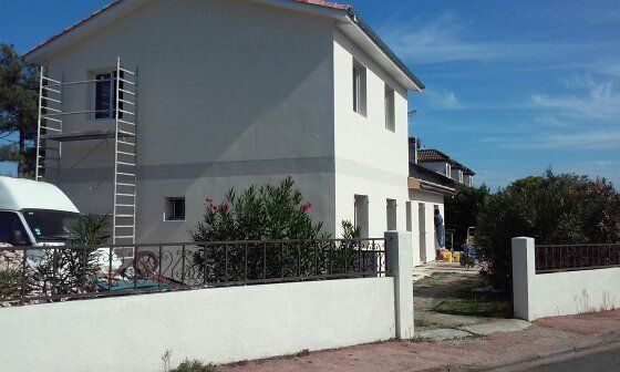 Une maison blanche de deux étages en rénovation, avec des échafaudages contre le mur et une camionnette garée dans la cour.